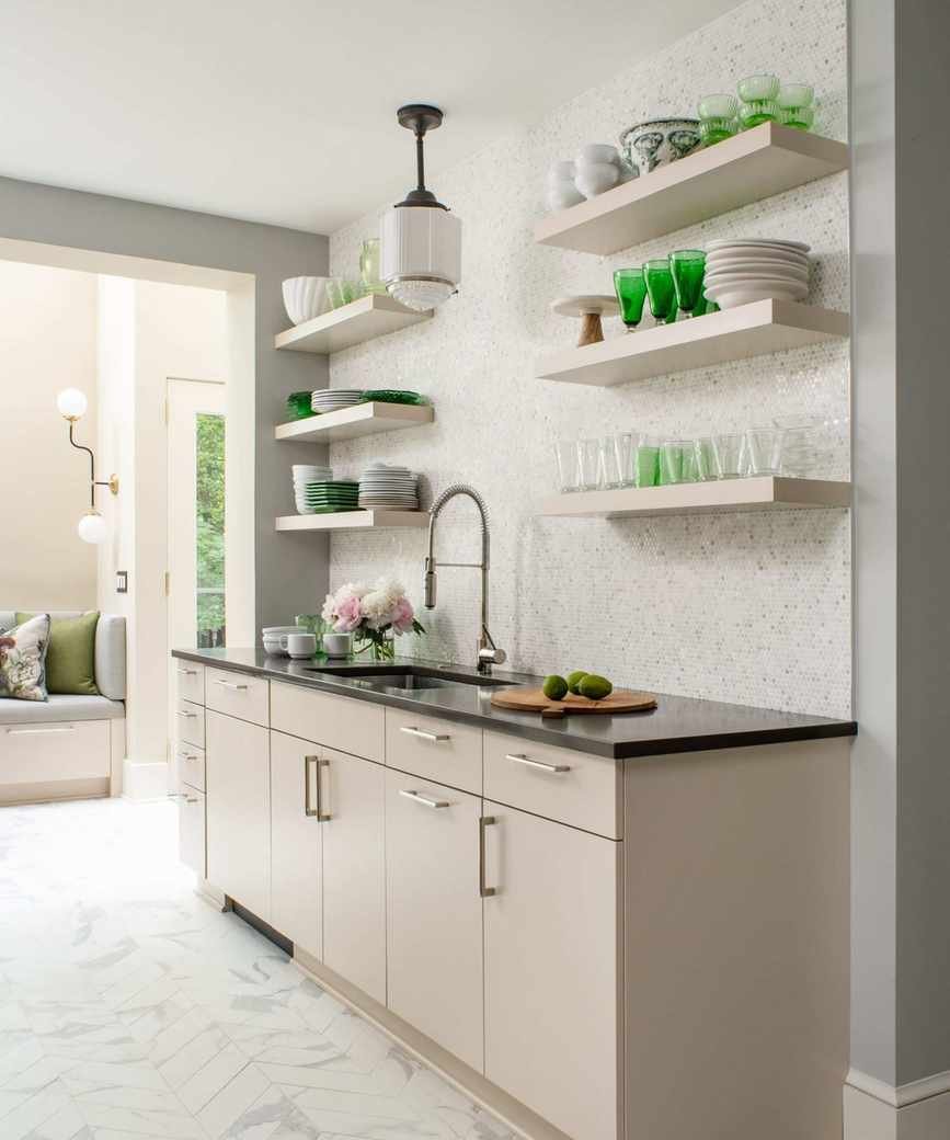 A neutral kitchen with white walls, wood cabinets, black countertops, and white floating wall shelves decorated with green glass decor and tableware