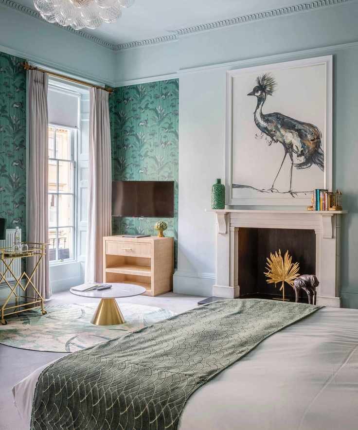 aqua/pale blue bedroom with large artwork above fireplace, drapes, throw on bed, coffee table, rug, wallpaper