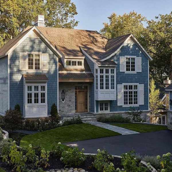 A home exterior with muted blue brickwork, white trim and window shutters, and a wooden front door. A front garden with lawn and plants, and a paved path.