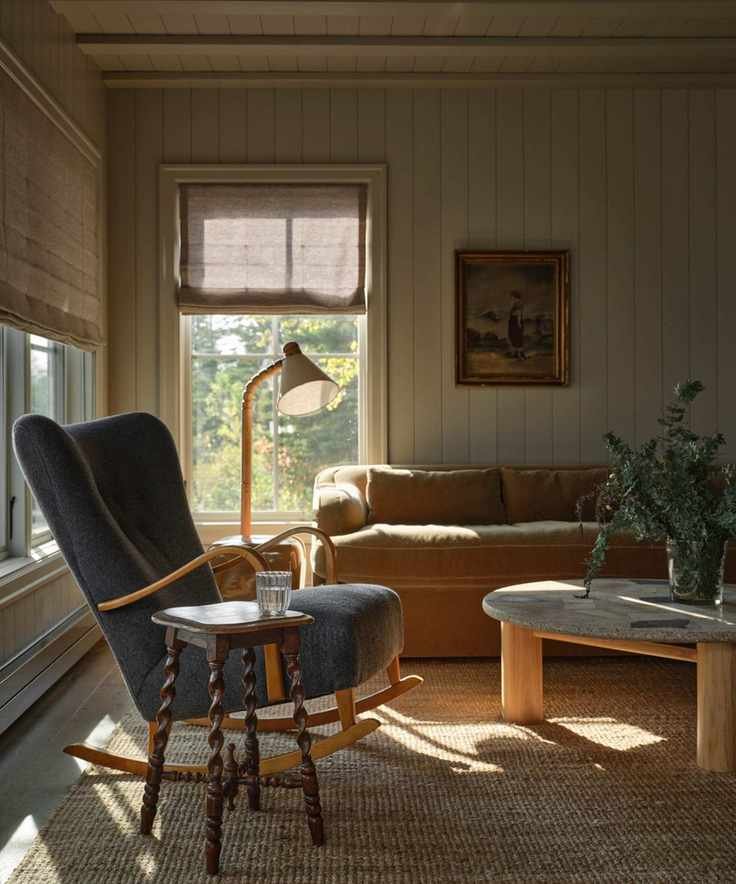 a vintage-styled paneled lakeside cottage living room with a beige velvet couch, vintage stone coffee table and a blue rocking chair