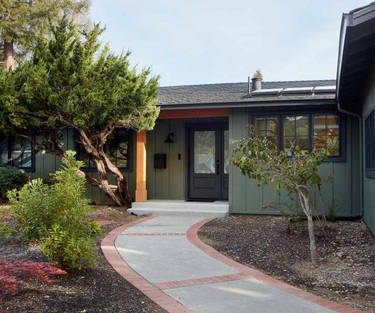 A bungalow home exterior with dark green siding, a charcoal gray front door and window trim, and an orange pillar. A paved pathway with surrounding plants.