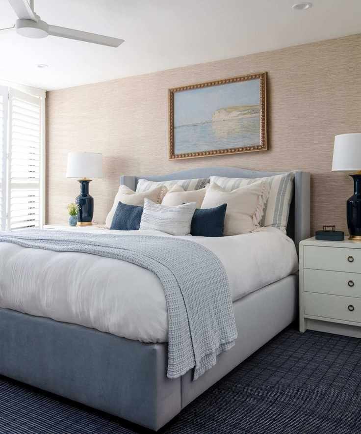 Coastal bedroom with grasscloth wall, white shutters, pale blue bed with blue and white bedding, blue rug and coastal painting