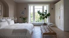 a cozy neutral bedroom with a large bed, a leather bench, a tree in front of the window, and a wall of cabinetry on the right side