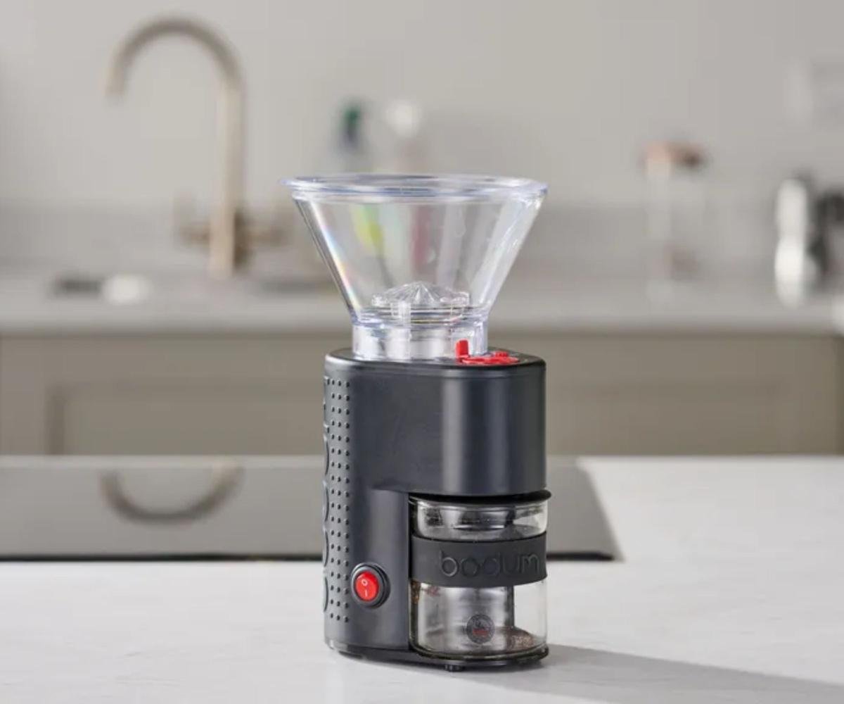 A compact black coffee grinder with a clear plastic funnel on the top, an out of focus kitchen in the background.