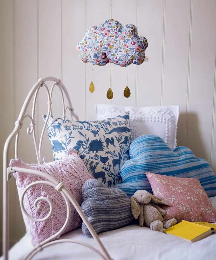Pastel pink and blue patterned and knitted cushions on a white child's daybed with hanging fabric cloud mobile.