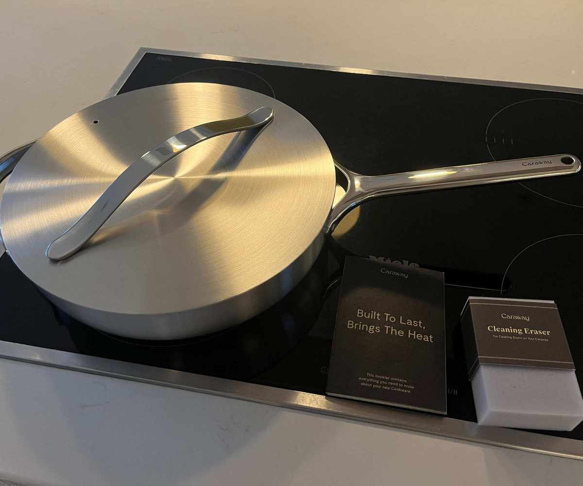 Caraway stainless steel sauté pan with a lid on a black electric stove top beside the instruction manual.