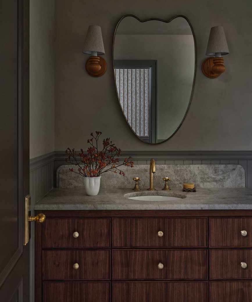 a cottage bathroom vanity area with dark brown wooden vanity, stone counter and a curved mirror with two wooden sconces either side