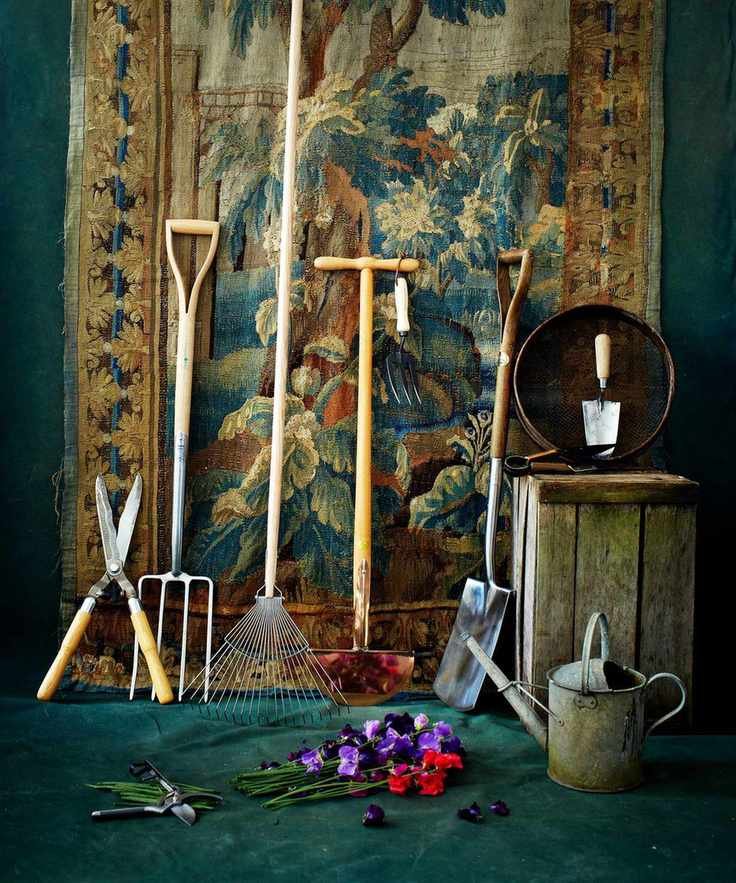 A tapestry wall hanging with an assortment of garden tools and a watering can leaning on it.