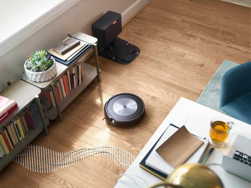 iRobot Roomba j7+ on wooden floor surrounded by furnishings: blue chair, bookshelf, plant, and table with notepads, laptop, and drink. The robot's clean base is against a wall behind the vacuum.