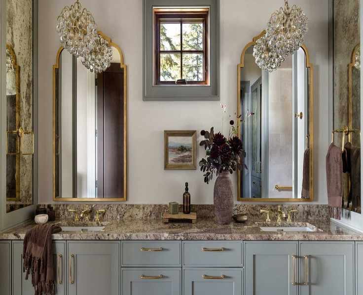 A sophisticated bathroom with warm white walls, pale blue cabinets, marble countertops, two brass mirrors on the wall, brass fixtures, and two glass pendant lights.