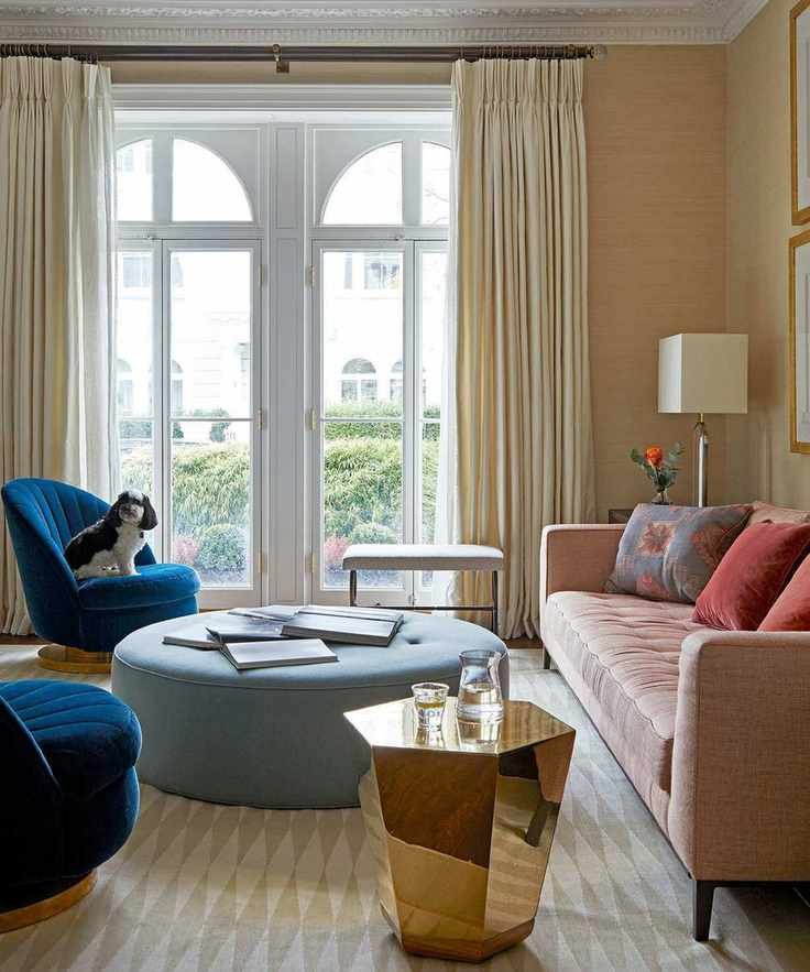 Living room with yellow textured wallpaper, cream diamond-pattern rug, pink sofa with colorful cushions, two deep blue lounge chairs, golden geometric side table, and a large rounded sky blue ottoman