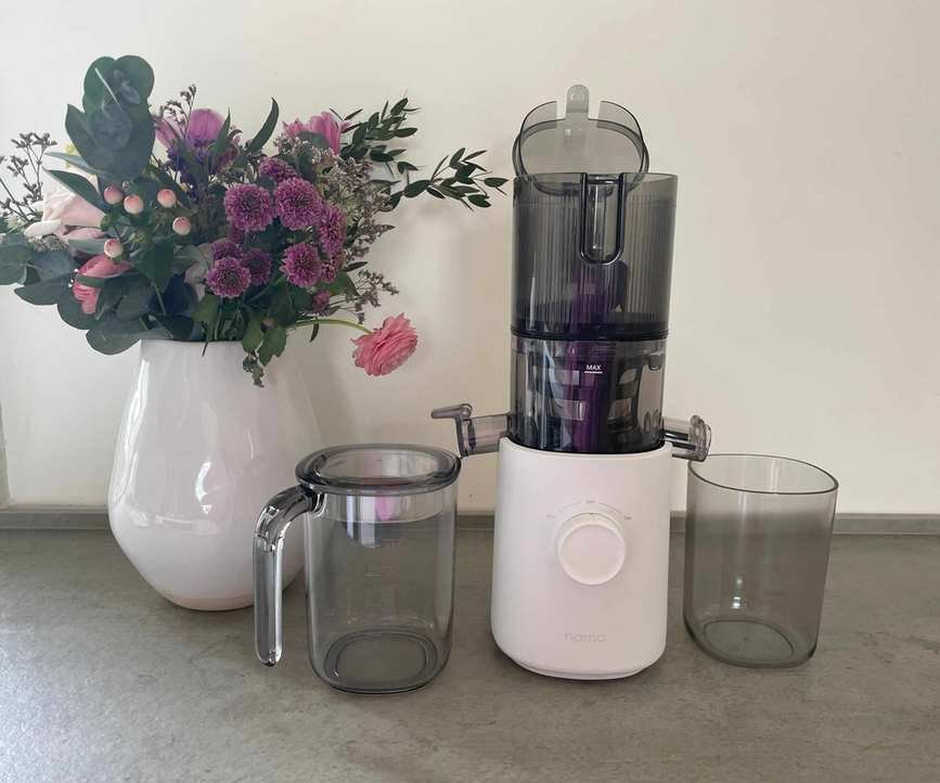 A cold press juicer with a white plastic base and a transparent grey plastic top, with lid is open. Two transparent grey plastic beakers sit either side.
