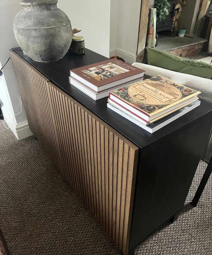 A black side table with fluted wooden front. Coffee table books stacked on top beside a large stone based lamp.