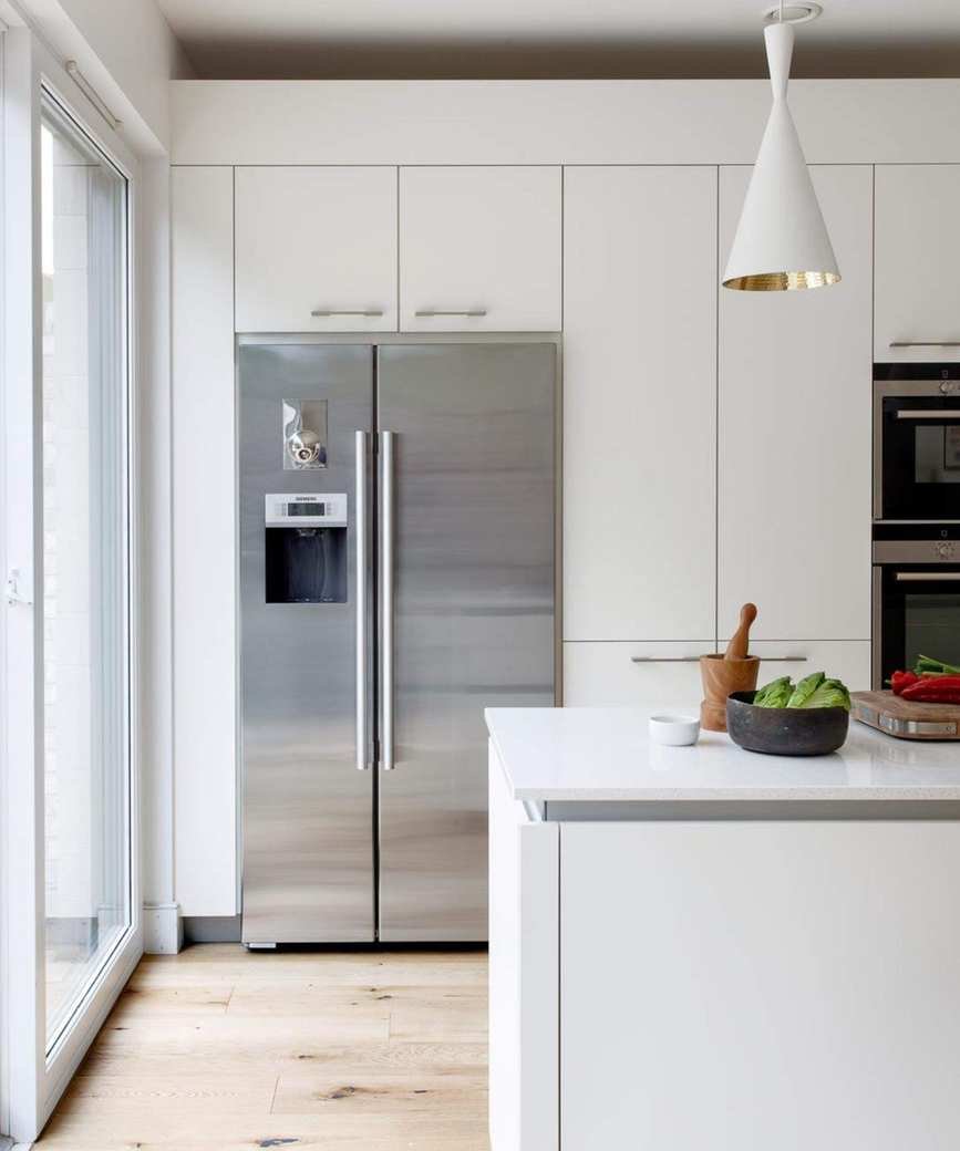 Stainless steel American style fridge freezer in a wall of floor-to-ceiling kitchen cabinets. The aesthetic of the kitchen is white, clean and minimalist with wooden floors and large kitchen island with spot lighting