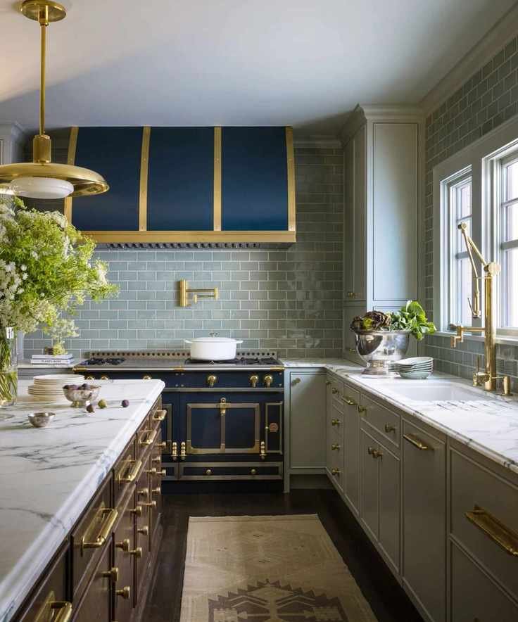 A large kitchen with light green cabinets, an island with a marble countertop, a dark blue oven and hood, pale green backsplash tiles, and a neutral runner rug on the dark flooring.