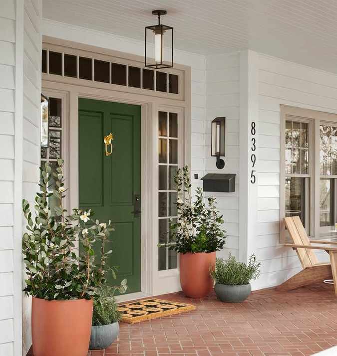 Traditional home front porch with green door, potted plants and a welcome mat. There is an Adirondack chair in view to the right and and the floor is red brick tile.