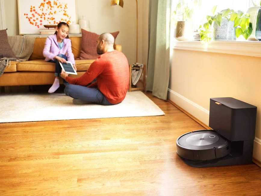 Parent sitting on a white rug interacting with a child on a brown sofa in the background, with the iRobot Roomba j7+ robot vacuum in the foreground on a wooden floor.