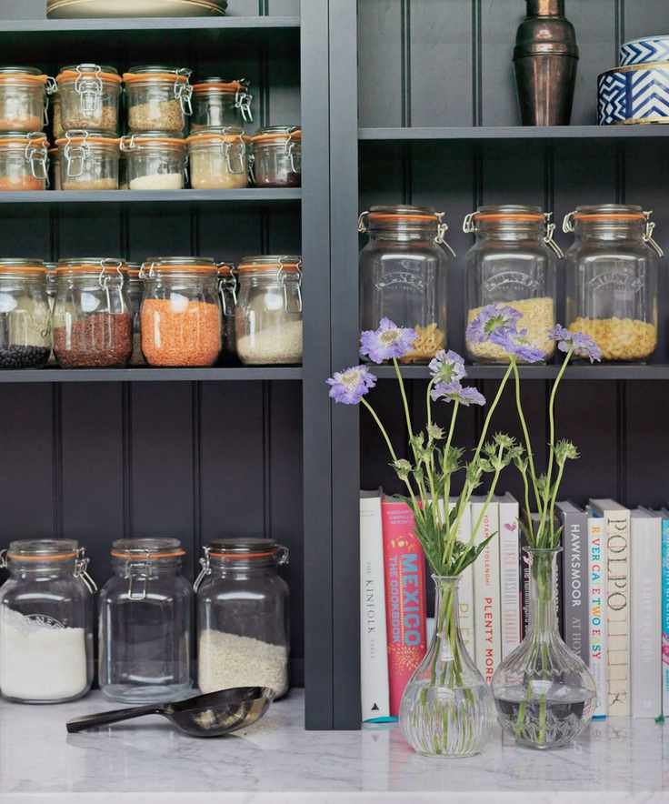 Dark gray pantry shelves with Kilner jars, recipe books and a copper cocktail shaker, with glass vases filled with purple flowers