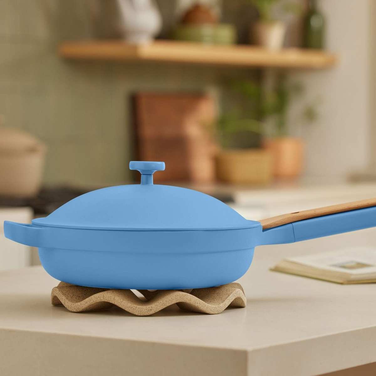 A blue pan with a long handle on one side and a carry handle opposite. A matching domed lid sits on top. The pan sits on a wavy trivet on a white quartz countertop, and there is an open cookbook out of focus to its right.