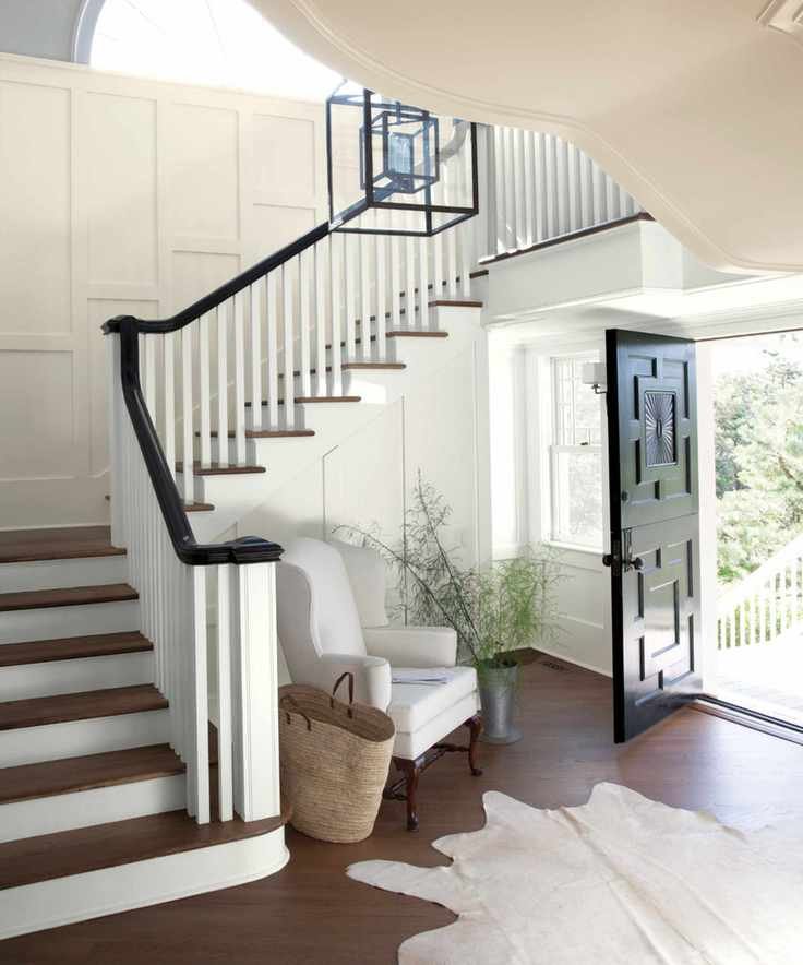 Large hallway with dark wooden flooring, white painted walls with wall paneling, large animal skin style rug, armchair near stairs, large stairway with black painted banister, dark wooden steps