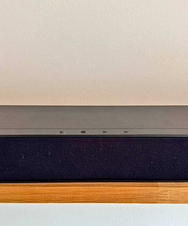 Closeup of Hisense HS2100 soundbar's four control buttons on wooden shelf in front of white wall.
