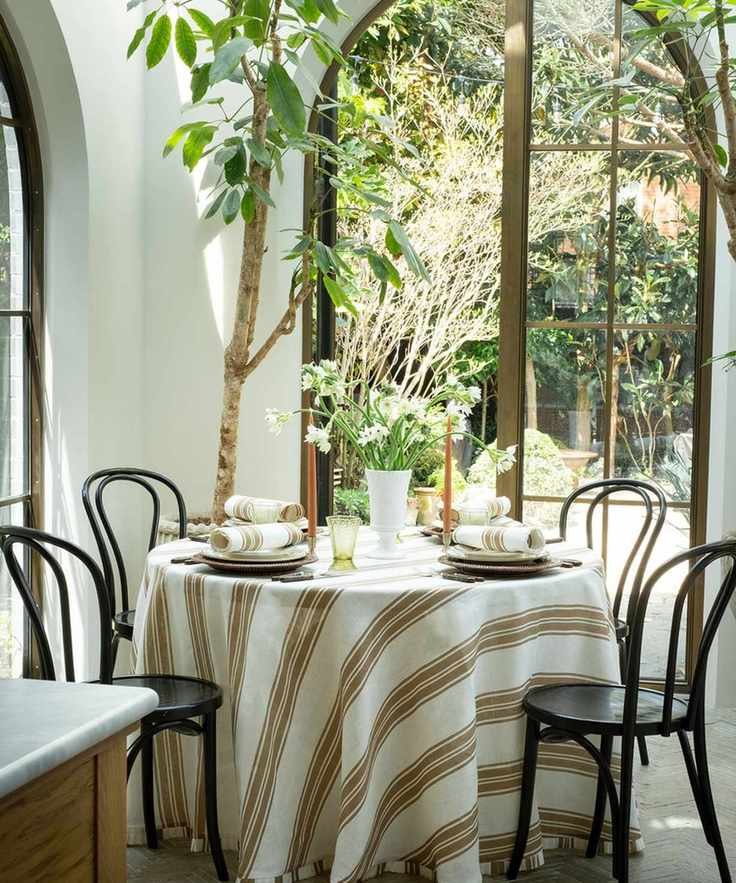indoor meets outdoor table set up in a garden room or sunroom with a round table, black wooden chairs, and a white and beige striped tablecloth