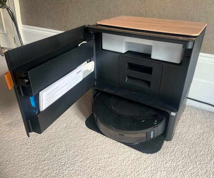 The view inside the clean base of the iRobot Roomba Combo j9+ in Alex's home. The user manual and robot vacuum are visible with two shelves and compartments for dust and water