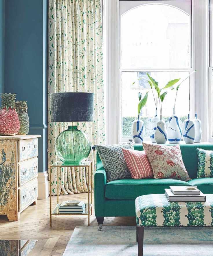 A bright and colorful living room with layered patterns, with a green sofa, a patterned rectangular foot rest, colorful cushions, a green glass lamp with a blue velvet shade, a patterned chest of drawers and large bay windows behind, with patterned curtains.