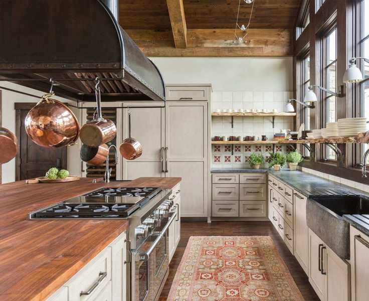 Large rustic kitchen, gray cabinetry, kitchen island with wooden countertop, pot rack hacking above, shelves decorated with kitchenware and glassware, rug on stone floor, wooden beamed ceiling