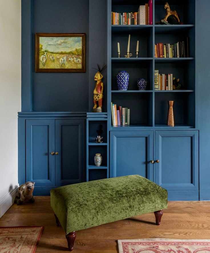 Dark blue painted storage unit with built-in shelving and books, dark wooden flooring with two rugs, and green fabric ottoman