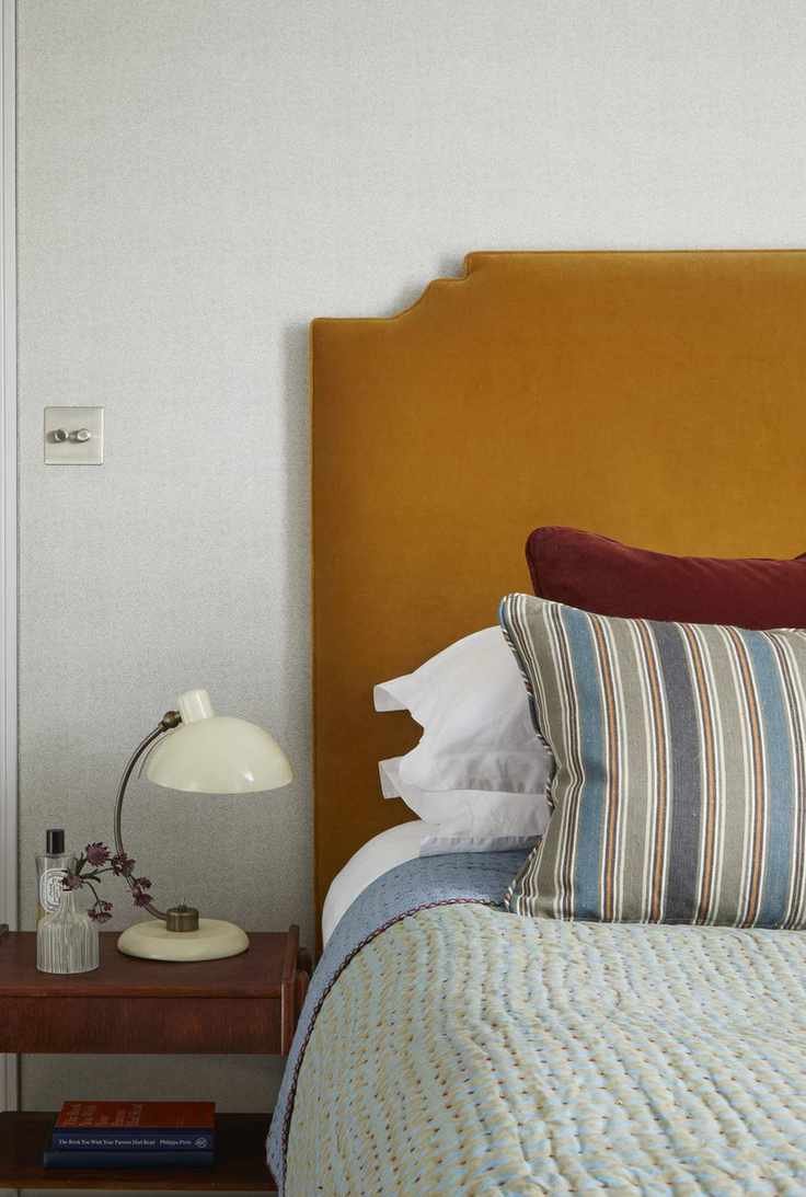 A bedroom with an orange velvet headboard, striped throw pillow and a wooden night stand with a white lamp