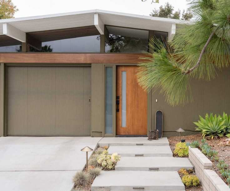 A modern home with olive green exterior, a sloped white roof, a glass upper wall, and a wooden front door with a modern front yard.