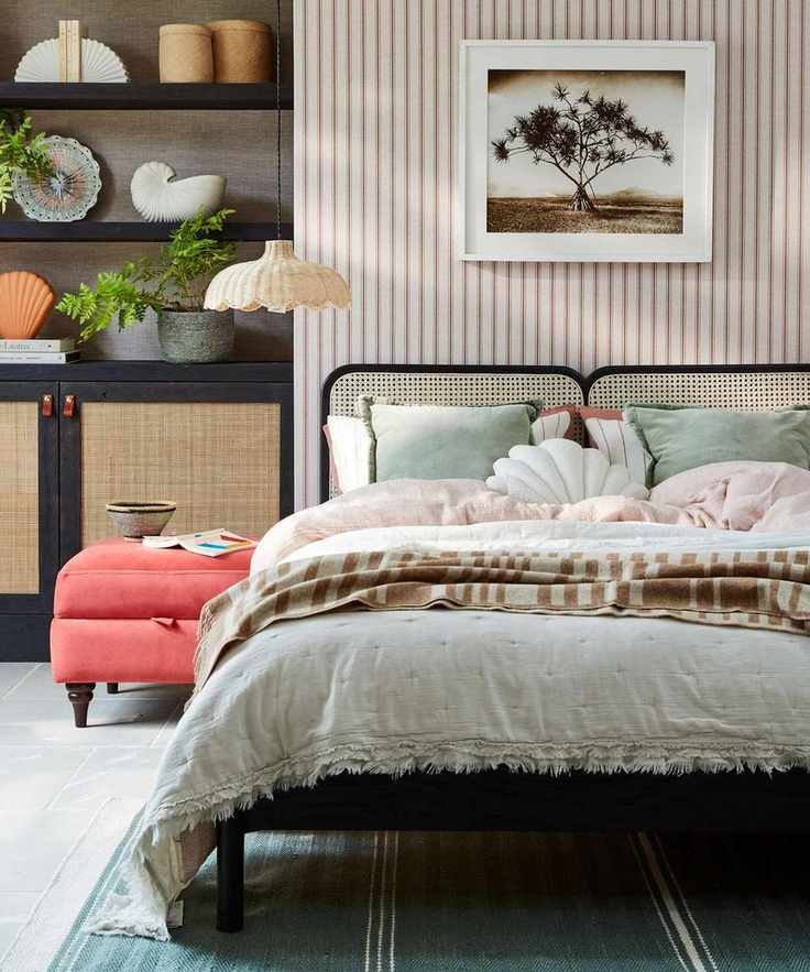 A bright and colorful bedroom with black shelves with plants and ornaments, striped wallpaper, rattan bed with colorful bedding and cushions, and a blue rug.