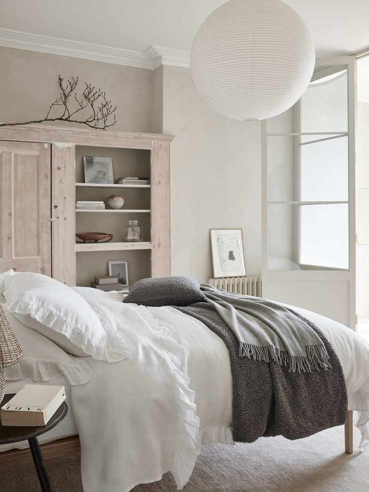 white and neutral bedroom with limed cabinet, stone walls, white bedding, cream carpet, gray blanket and cushion, white paper lampshade