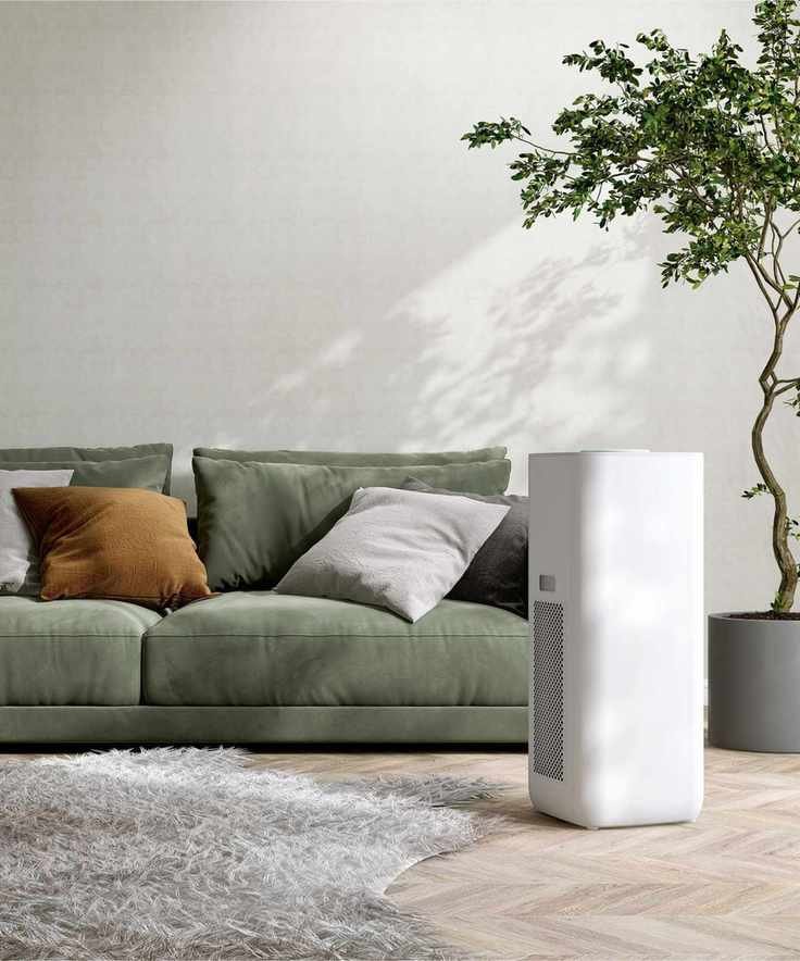 A white air purifier beside a green sofa and indoor tree, with a white wall and wooden flooring