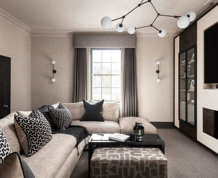 Modern gray living room with gray curtains and pelmet on large window, gray corner sofa, gray painted walls, gray patterned ottoman and black table in front of sofa, black media unit with TV, matching ceiling and wall lights