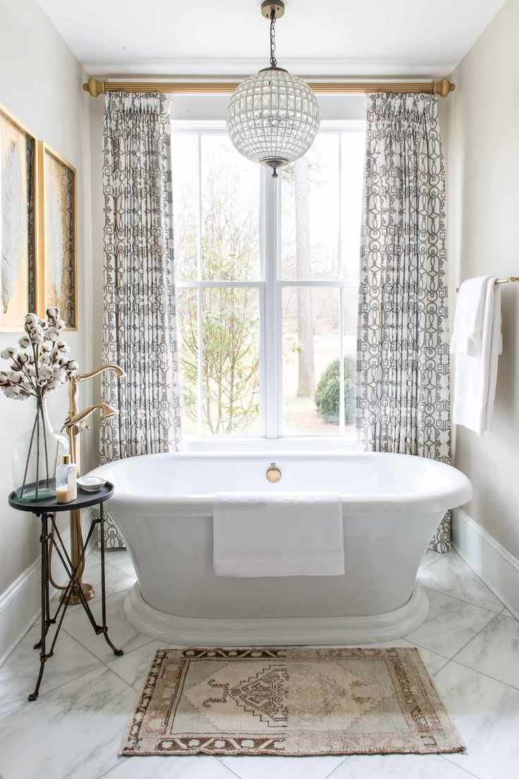 neutral bathroom with white roll top bath, chandelier, marble floor tiles black and white curtains