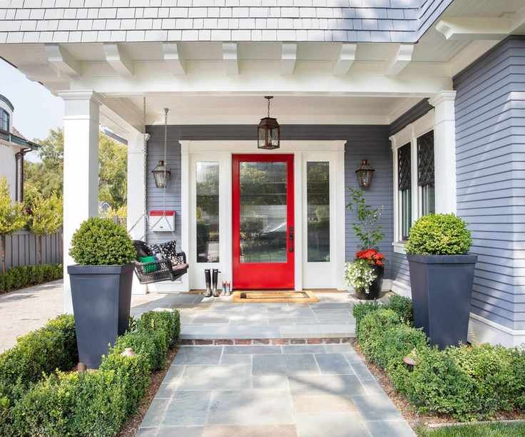 A home exterior painted gray with white trim and a red front door with a glass panel. A porch with lantern-style wall and pendant lights, and potted plants.