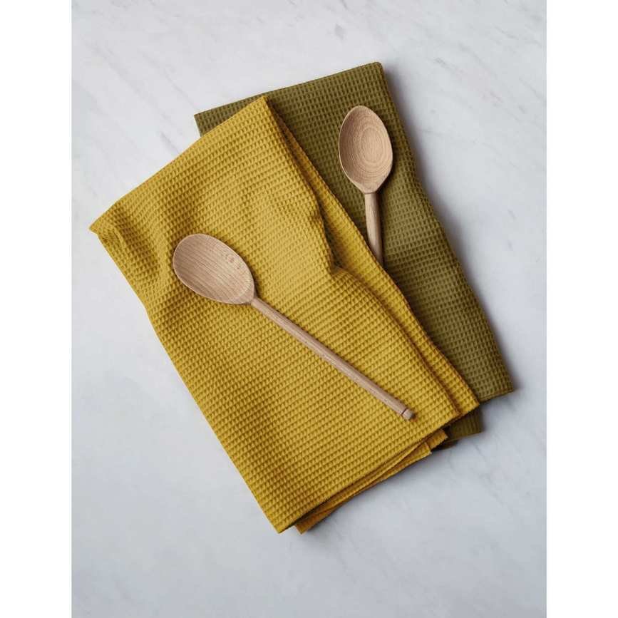 Two waffle cotton tea towels, one yellow one green, on top of one another. Wooden spoons on top of each towel