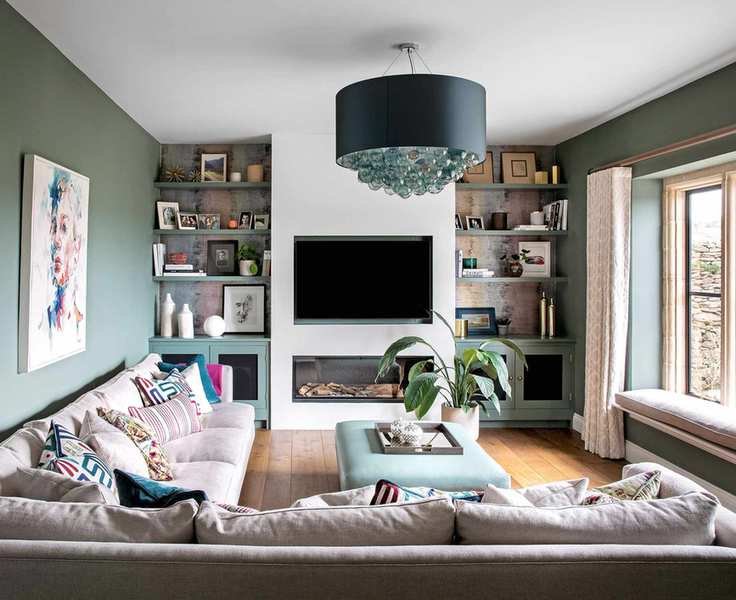 Family room with green walls, large cream sectional sofa adorned with colorful cushions, light green ottoman, dark wood floor, white-painted chimney breast with wall-mounted TV and fireplace, built-in shelves with decorative items, illuminated by a large green pendant light, and colorful artwork, featuring window bench seating with cream cushions