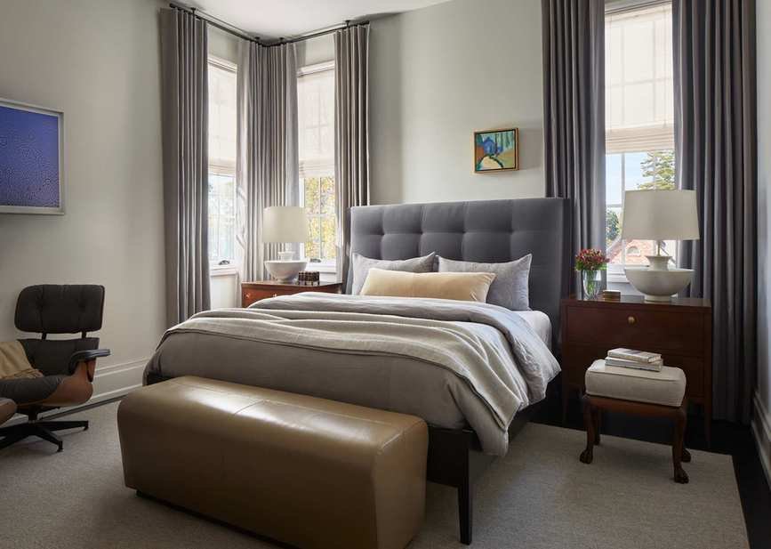 A Mid-Century Modern style bedroom with a light gray studded headboard, gray curtains in the windows, and a leather bench at the foot of the bed