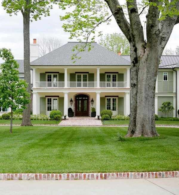 A traditional-style house exterior with muted green and soft white paintwork, a lush green lawn with trees, and a paved path.