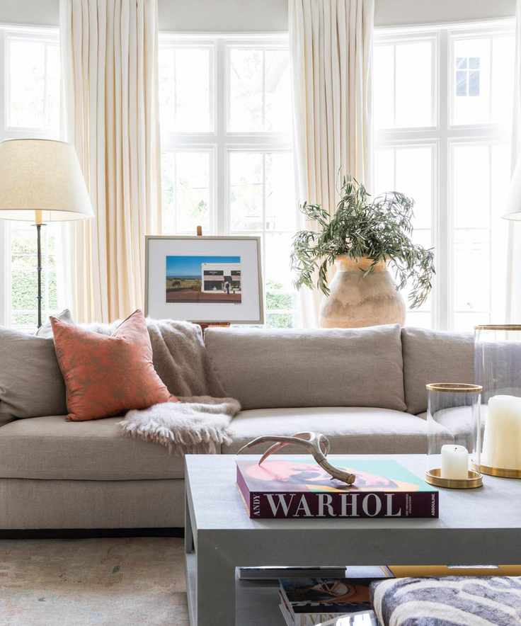 living room with cream drapes, gray couch, coffee table with books and candles, rust couch pillows, floor lamps