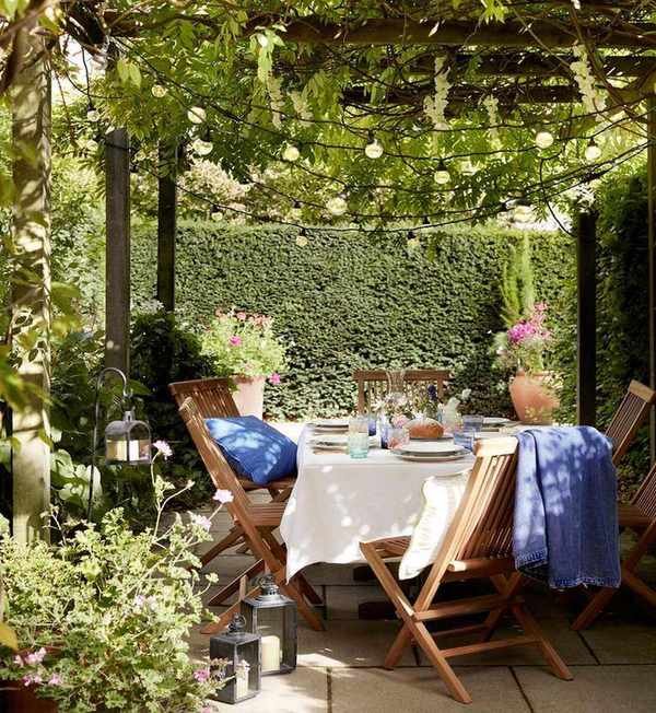 Wooden dining setup in a small garden under a pergola with lights.