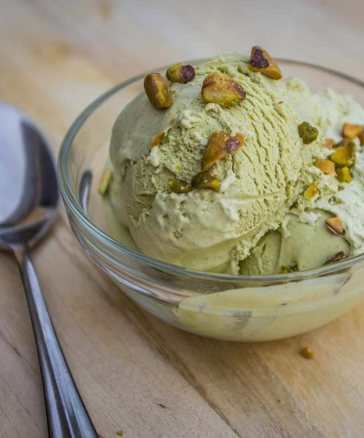 Glass bowl of pistachio ice cream with crushed pistachios on top, placed on a wooden table with a silver spoon beside it
