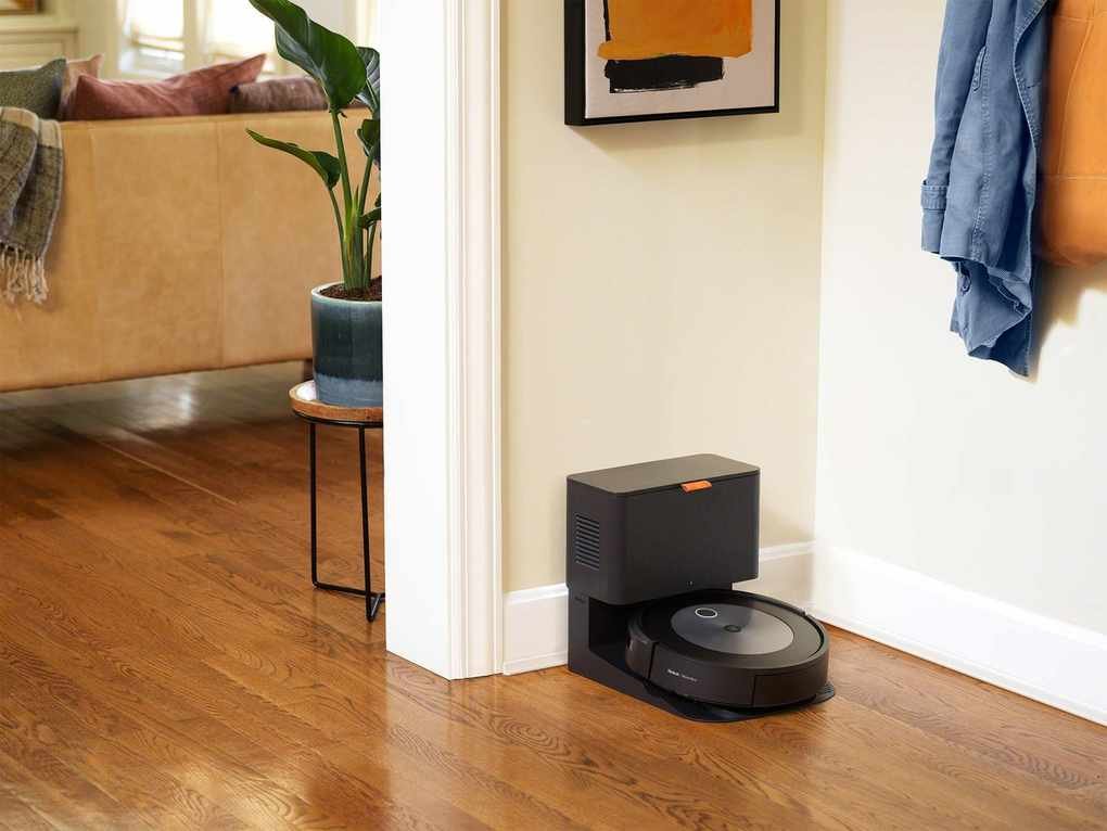 iRobot Roomba j7+ docked on charging base against white wall, on wooden floor with living room furniture visible in the background.