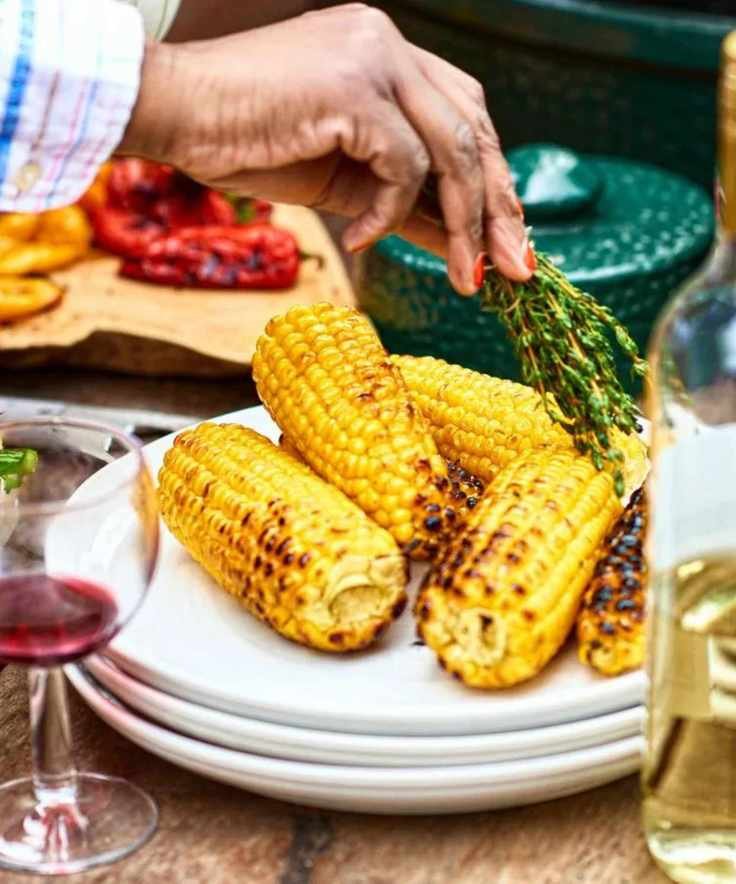 Grilled corn on a plate at a party with a glass of wine, a bottle of wine, being brushed with rosemary