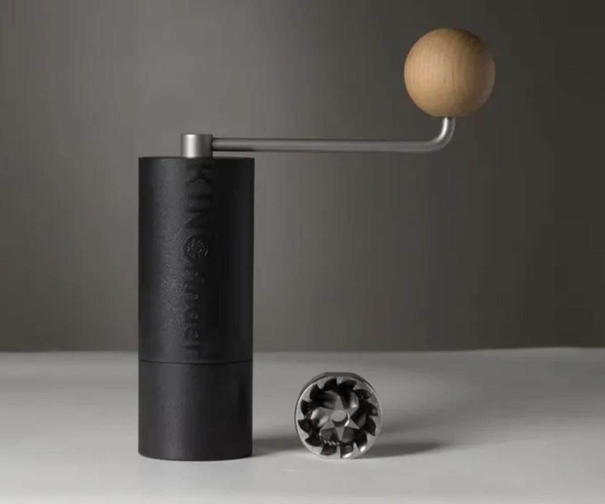 A black cylindrical coffee grinder on a white countertop, with a stainless steel crank handle.