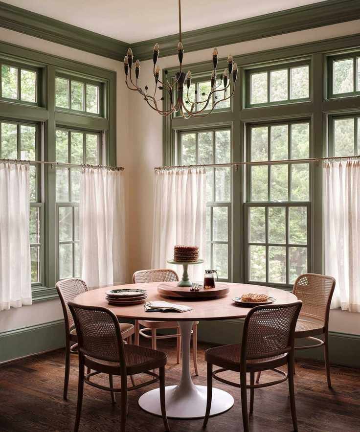 A breakfast nook in a kitchen with large windows with green woodwork and cafe curtains, wood floors, a round dining table, and wooden chairs.
