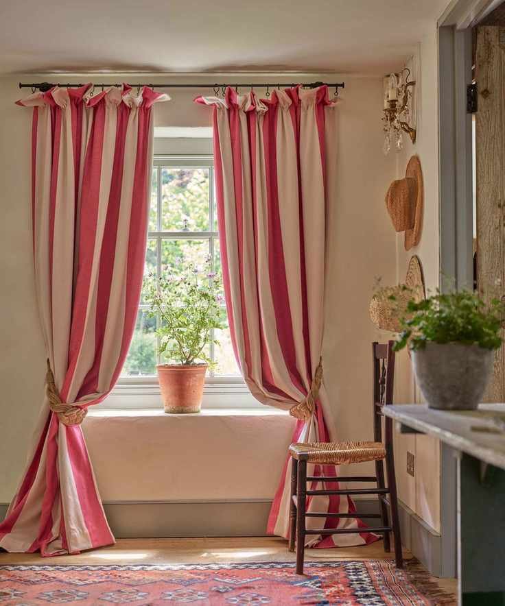 Country style living room with red and white striped curtains tied back on window, white painted walls, gray skirting board, light wood flooring, red patterned rug